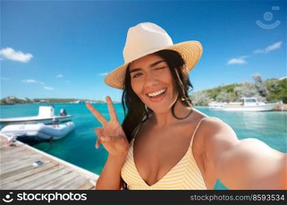 people, summer and swimwear concept - happy smiling woman in bikini swimsuit and straw hat taking selfie and showing peace gesture over pier and boat or tropical sea background in french polynesia. smiling woman in bikini taking selfie on pier