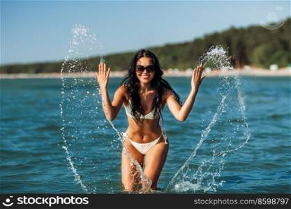 people, summer and swimwear concept - happy smiling woman in bikini swimsuit splashing sea water on beach. woman in bikini splashing sea water on beach