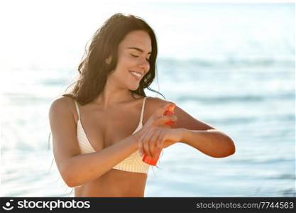 people, summer and swimwear concept - happy smiling young woman in bikini swimsuit using sunscreen spray on beach. smiling woman in bikini with sunscreen on beach