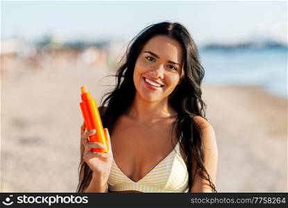 people, summer and swimwear concept - happy smiling young woman in bikini swimsuit with sunscreen spray on beach. smiling woman in bikini with sunscreen on beach