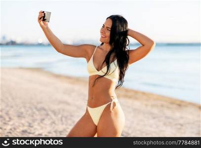 people, summer and swimwear concept - happy smiling young woman in bikini swimsuit taking selfie with smartphone on beach. smiling woman in bikini taking selfie on beach