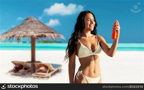 people, summer and swimwear concept - happy smiling young woman in bikini swimsuit using sunscreen spray over palapa and sun beds on tropical beach background in french polynesia. smiling woman in bikini with sunscreen on beach