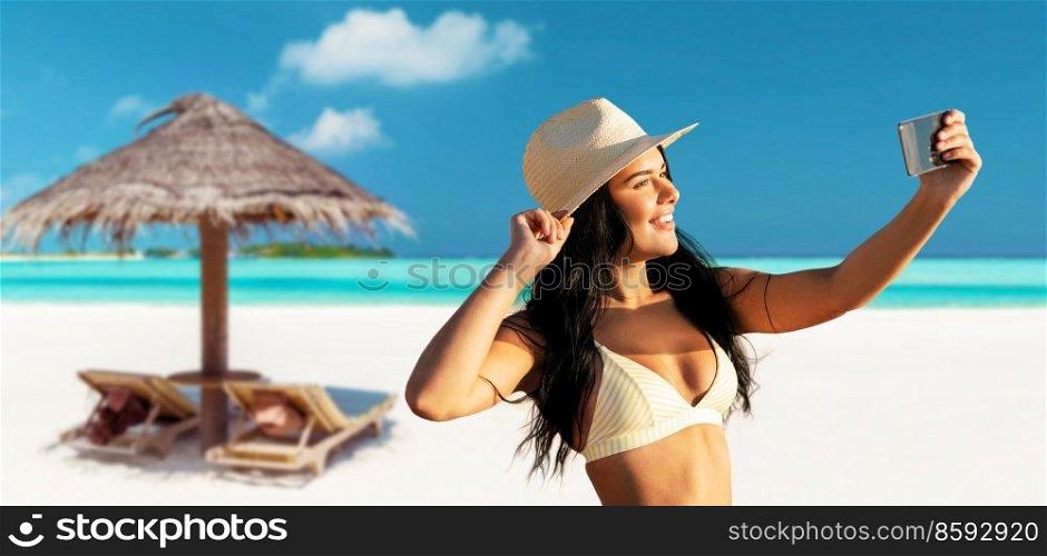 people, summer and swimwear concept - happy smiling young woman in bikini swimsuit and straw hat taking selfie with smartphone over palapa and sun beds on tropical beach background in french polynesia. smiling woman in bikini taking selfie on beach