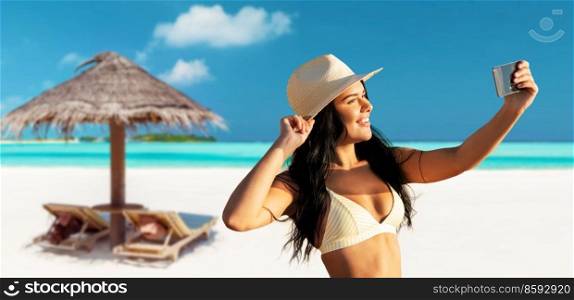 people, summer and swimwear concept - happy smiling young woman in bikini swimsuit and straw hat taking selfie with smartphone over palapa and sun beds on tropical beach background in french polynesia. smiling woman in bikini taking selfie on beach