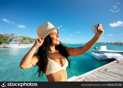people, summer and swimwear concept - happy smiling young woman in bikini swimsuit and straw hat taking selfie with smartphone over pier and boat or tropical sea background in french polynesia. smiling woman in bikini taking selfie on pier