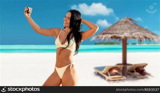 people, summer and swimwear concept - happy smiling young woman in bikini swimsuit taking selfie with smartphone over palapa and sun beds on tropical beach background in french polynesia. smiling woman in bikini taking selfie on beach