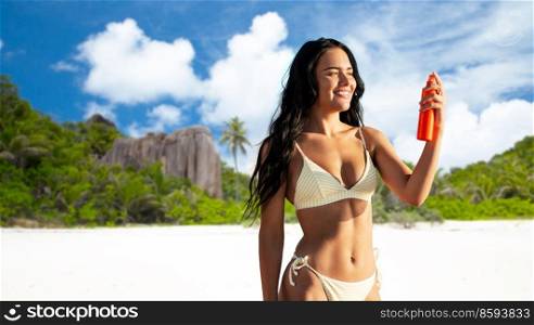 people, summer and swimwear concept - happy smiling young woman in bikini swimsuit using sunscreen spray over seychelles island beach background. smiling woman in bikini with sunscreen on beach