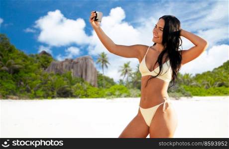 people, summer and swimwear concept - happy smiling young woman in bikini swimsuit taking selfie with smartphone over seychelles island beach background. smiling woman in bikini taking selfie on beach