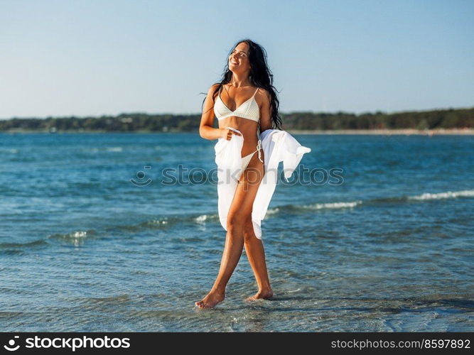 people, summer and swimwear concept - happy smiling young woman in bikini swimsuit with pareo on beach. woman in bikini swimsuit with pareo on beach