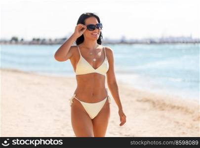 people, summer and swimwear concept - happy smiling young woman in bikini swimsuit and sunglasses on beach. smiling young woman in bikini swimsuit on beach