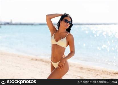 people, summer and swimwear concept - happy smiling young woman in bikini swimsuit and sunglasses posing on beach. smiling young woman in bikini swimsuit on beach