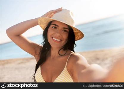 people, summer and swimwear concept - happy smiling young woman in bikini swimsuit and straw hat taking selfie on beach. smiling woman in bikini taking selfie on beach