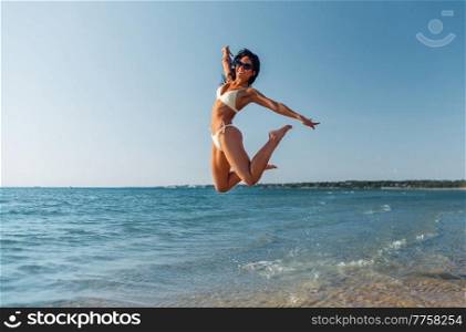 people, summer and swimwear concept - happy smiling young woman in bikini swimsuit jumping on beach. smiling young woman in bikini swimsuit on beach