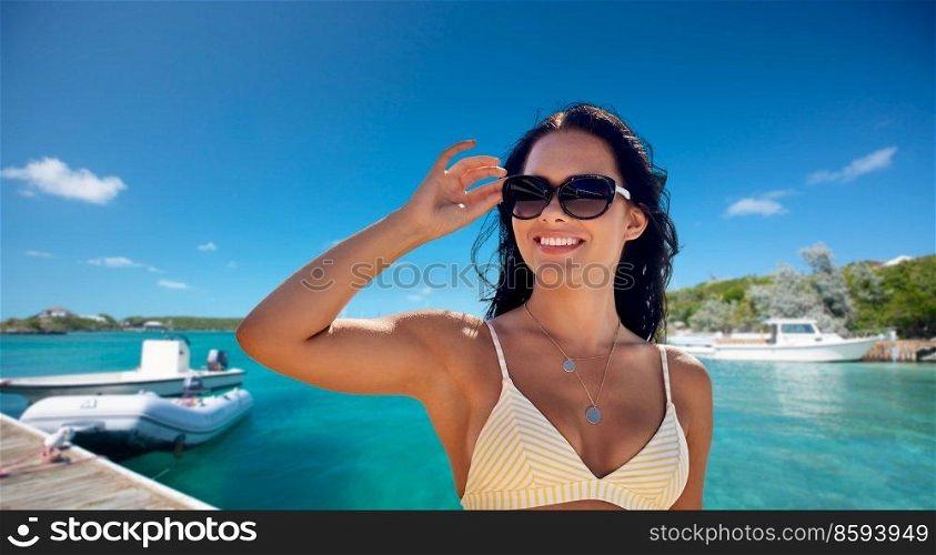 people, summer and swimwear concept - happy smiling young woman in sunglasses and bikini swimsuit over pier and boat or tropical sea background in french polynesia. smiling woman in sunglasses on pier in summer