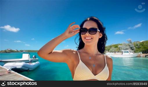 people, summer and swimwear concept - happy smiling young woman in sunglasses and bikini swimsuit over pier and boat or tropical sea background in french polynesia. smiling woman in sunglasses on pier in summer