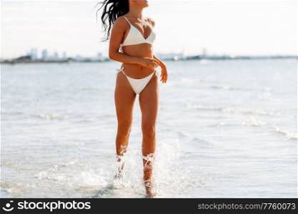 people, summer and swimwear concept - happy young woman in bikini swimsuit running in water beach. young woman in bikini swimsuit on beach