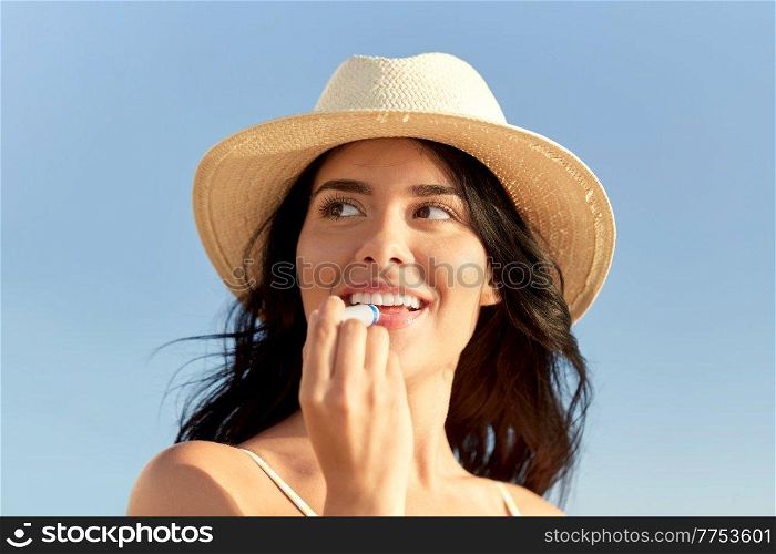 people, summer and swimwear concept - portrait of happy smiling young woman in bikini swimsuit and straw hat applying lip balm on beach. smiling woman in bikini with lip balm on beach