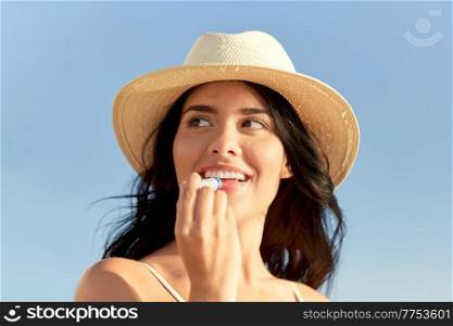 people, summer and swimwear concept - portrait of happy smiling young woman in bikini swimsuit and straw hat applying lip balm on beach. smiling woman in bikini with lip balm on beach