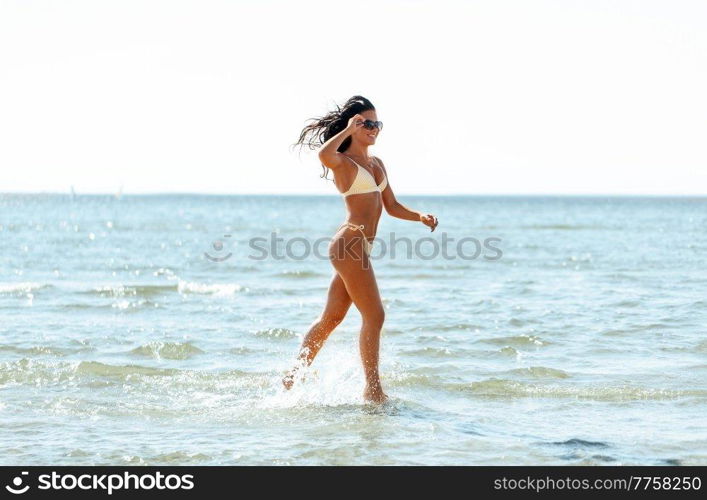 people, summer and swimwear concept - young woman in bikini swimsuit running in shallow water on beach. young woman in bikini swimsuit running on beach