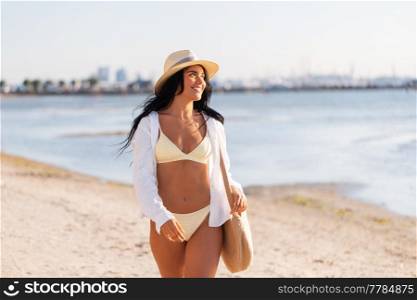 people, summer holidays and leisure concept - happy young woman in bikini swimsuit, white shirt and straw hat with bag walking along beach. happy woman in bikini and shirt walking on beach