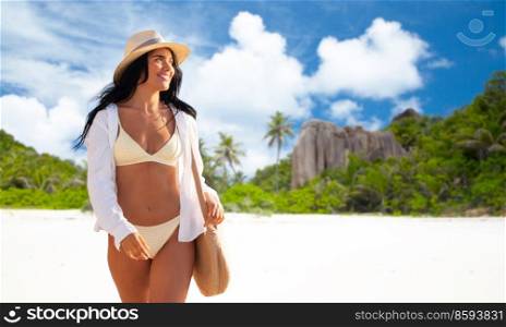 people, summer holidays and leisure concept - happy young woman in bikini swimsuit, white shirt and straw hat with bag over seychelles island beach background. happy woman in bikini and shirt walking on beach