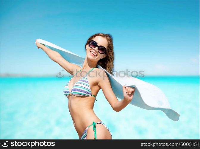 people, summer holidays and vacation concept - beautiful woman in bikini and sunglasses with towel on beach over blue sky and sea background. woman in bikini and sunglasses with towel on beach