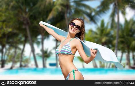 people, summer holidays and vacation concept - beautiful woman in bikini and sunglasses with towel over exotic tropical beach with palm trees and pool background. woman in bikini and sunglasses with towel on beach
