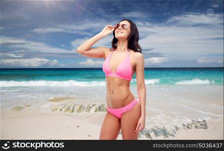people, summer holidays, travel and tourism concept - happy young woman in sunglasses and pink swimsuit looking up over exotic tropical beach and ocean background. happy woman in sunglasses and bikini on beach
