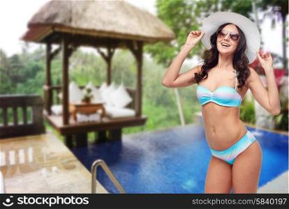 people, summer holidays, travel and tourism concept - happy young woman in bikini swimsuit, sunglasses and sun hat over hotel resort background