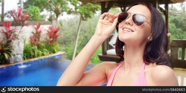 people, summer holidays, travel and tourism concept - happy young woman in sunglasses and pink swimsuit looking up over hotel resort background