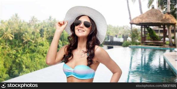 people, summer holidays, travel and tourism concept - happy young woman in bikini swimsuit, sunglasses and sun hat over infinity edge swimming pool background