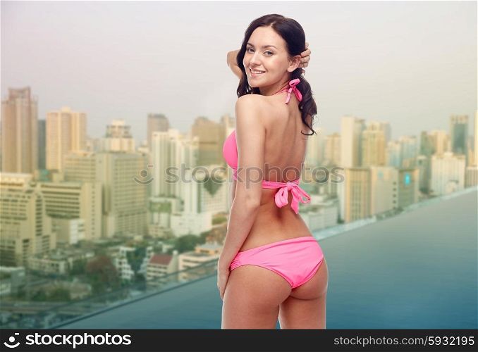 people, summer holidays, travel and tourism concept - happy young woman in pink bikini swimsuit looking back over infinity edge swimming pool and city background