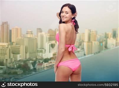 people, summer holidays, travel and tourism concept - happy young woman in pink bikini swimsuit looking back over infinity edge swimming pool and city background