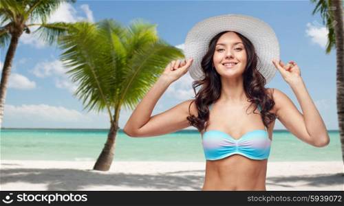 people, summer holidays, travel and tourism concept - happy young woman in bikini swimsuit and sun hat over tropical beach with palm trees background