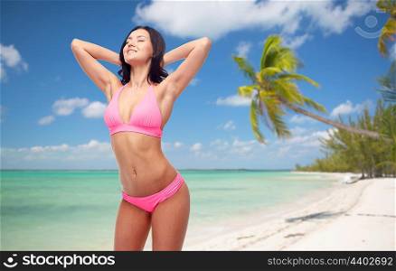 people, summer holidays, travel and tourism concept - happy young woman posing in pink bikini swimsuit over exotic tropical beach with palm trees background,