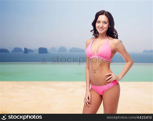 people, summer holidays, travel and tourism concept - happy young woman posing in pink bikini swimsuit over beach or infinity edge pool and ocean background