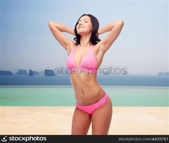 people, summer holidays, travel and tourism concept - happy young woman posing in pink bikini swimsuit over beach or infinity edge pool and ocean background