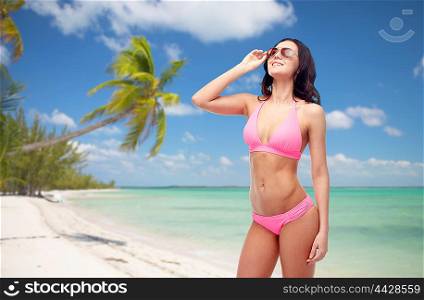 people, summer holidays, travel and tourism concept - happy young woman posing in pink bikini swimsuit and sunglasses over exotic tropical beach with palm trees background