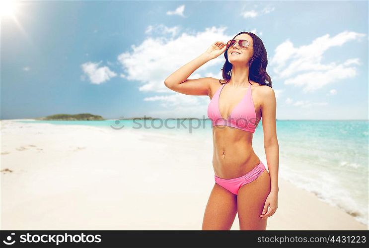 people, summer holidays, travel and tourism concept - happy young woman posing in pink bikini swimsuit and sunglasses over exotic tropical beach background