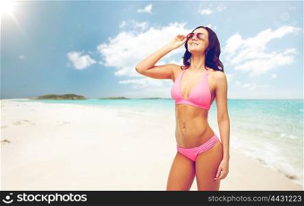 people, summer holidays, travel and tourism concept - happy young woman posing in pink bikini swimsuit and sunglasses over exotic tropical beach background