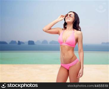 people, summer holidays, travel and tourism concept - happy young woman posing in pink bikini swimsuit and sunglasses over beach or infinity edge pool and ocean background