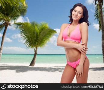 people, summer holidays, travel and tourism concept - happy young woman posing in pink bikini swimsuit over tropical beach with palm trees background