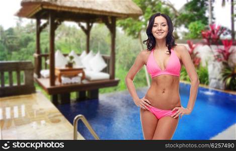 people, summer holidays, travel and tourism concept - happy young woman posing in pink bikini swimsuit over hotel resort background