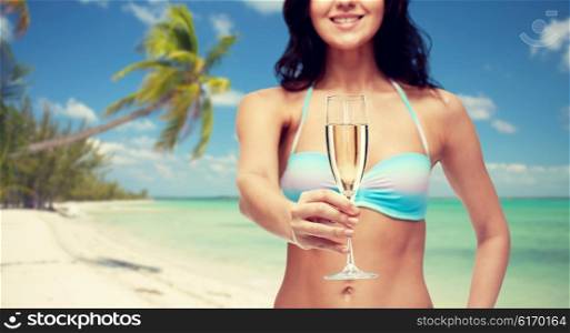 people, summer holidays, travel, celebration and drinks concept - happy young woman in bikini swimsuit drinking champagne at party over tropical beach background