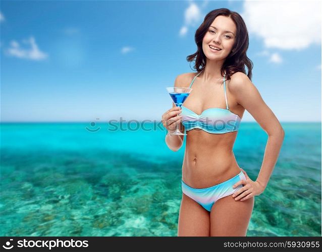 people, summer holidays, travel, tourism and alcohol drinks concept - happy young woman in bikini swimsuit holding glass of cocktail at beach party over sea and blue sky background