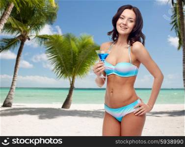 people, summer holidays, travel, tourism and alcohol drinks concept - happy young woman in bikini swimsuit holding glass of cocktail at party over tropical beach with palm trees background