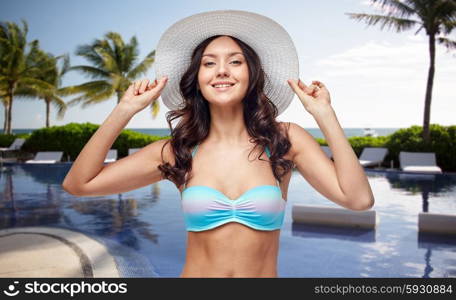 people, summer holidays, travel, tourism and beach concept - happy young woman in bikini swimsuit and sun hat over swimming pool with palm trees background