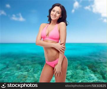 people, summer holidays, travel, tourism and beach concept - happy young woman posing in pink bikini swimsuit over sea and blue sky background