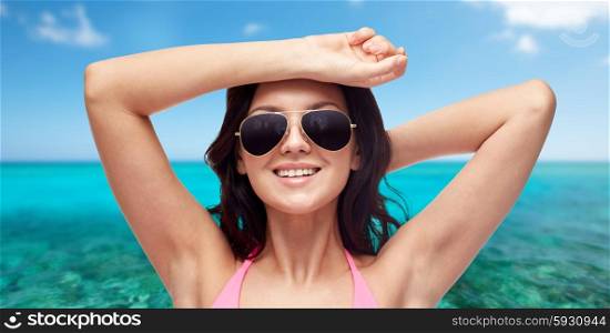 people, summer holidays, travel, tourism and beach concept - happy young woman in sunglasses and pink swimsuit over sea and blue sky background