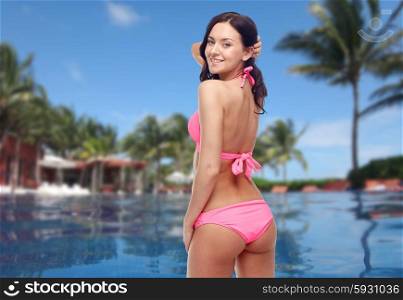 people, summer holidays, travel, tourism and beach concept - happy young woman in pink bikini swimsuit looking back over swimming pool with palm trees background